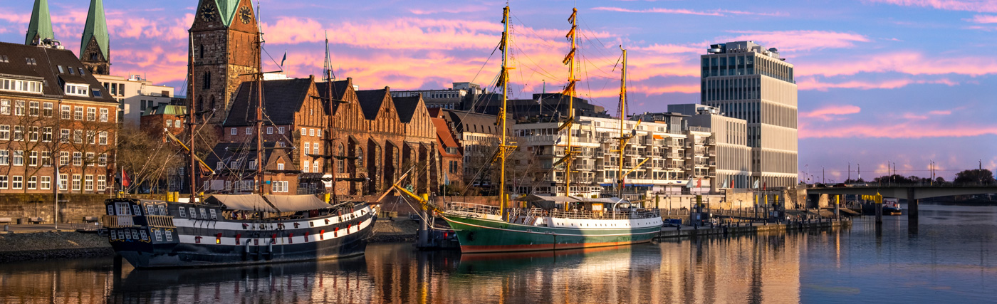 Zwei Großsegler liegen bei historischen und modernen Gebäuden entlang einer ruhigen Uferpromenade in Bremen vor Anker, während im Hintergrund ein farbenfroher Himmel beim Sonnenuntergang zu sehen ist.