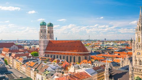 Panoramic view of Munich cityscape with the Frauenkirche and New Town Hall under a blue sky with scattered clouds.
