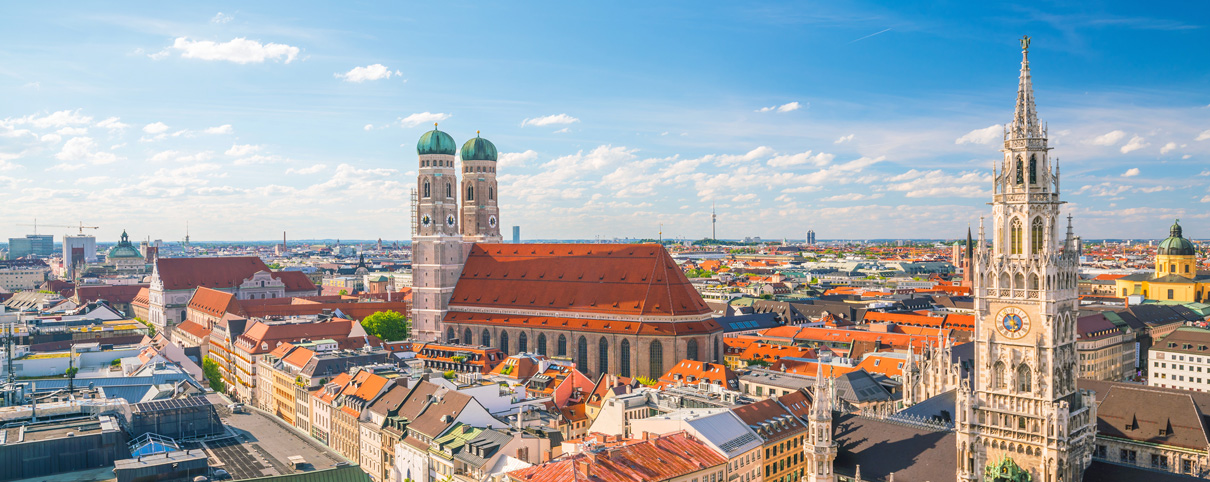Panoramic view of Munich cityscape with the Frauenkirche and New Town Hall under a blue sky with scattered clouds.