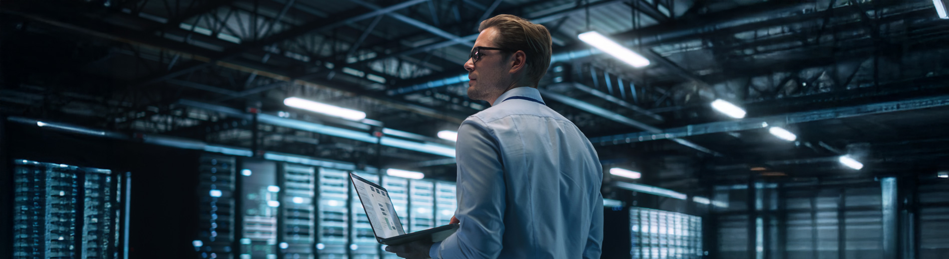 Mann mit Brille und Hemd hält Laptop in einem großen, modernen Serverraum mit Reihen von Serverracks